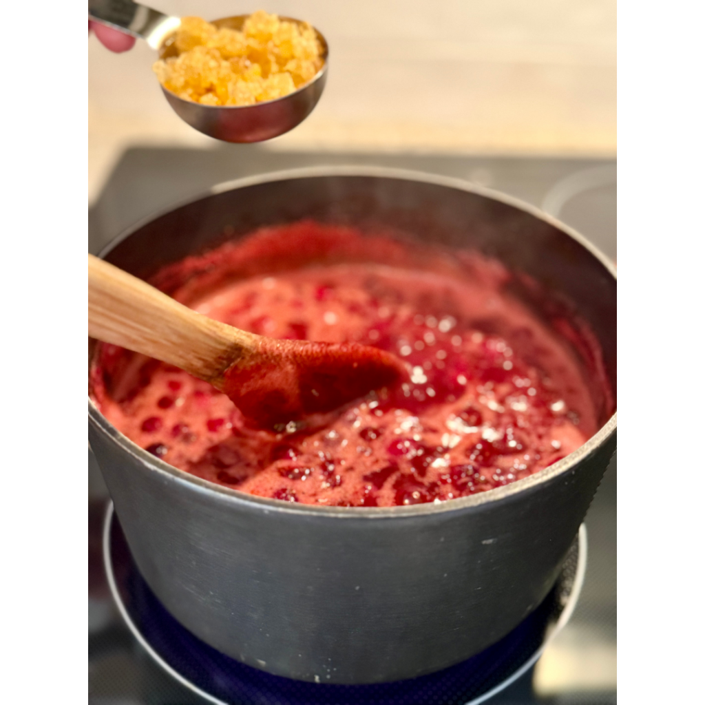 Cranberry sauce in medium sauce pan on stove top while woman adds chopped crystalized ginger into cranberry sauce.