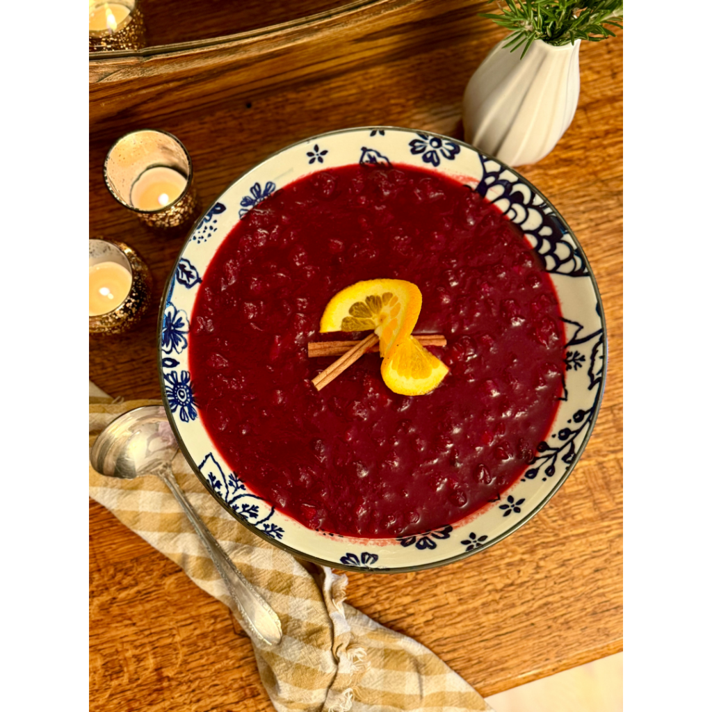 Cranberry sauce in blue and white serving bowl on wooden hutch with candles, gingham cloth napkin, serving spoon, and vase of rosemary.