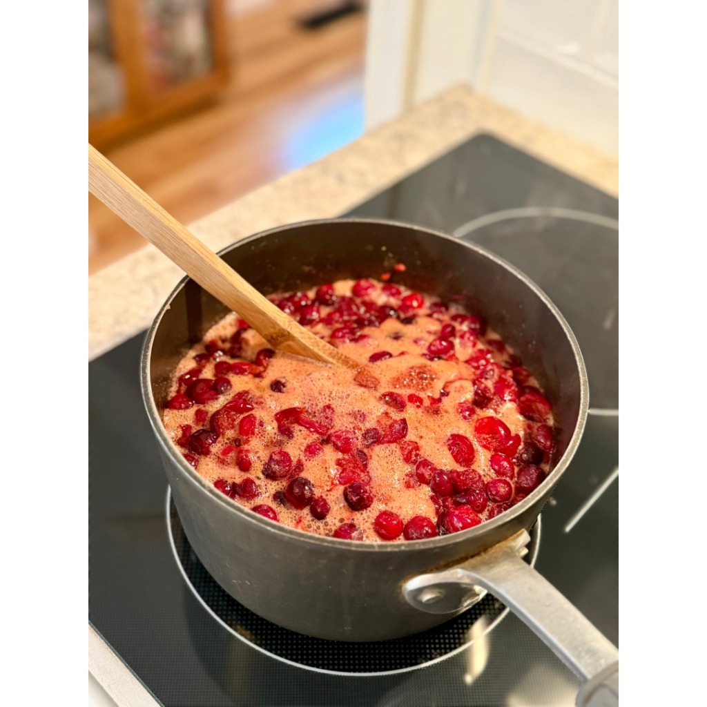 Cranberries in medium sauce pan at a light boil on stovetop with wooden spoon.