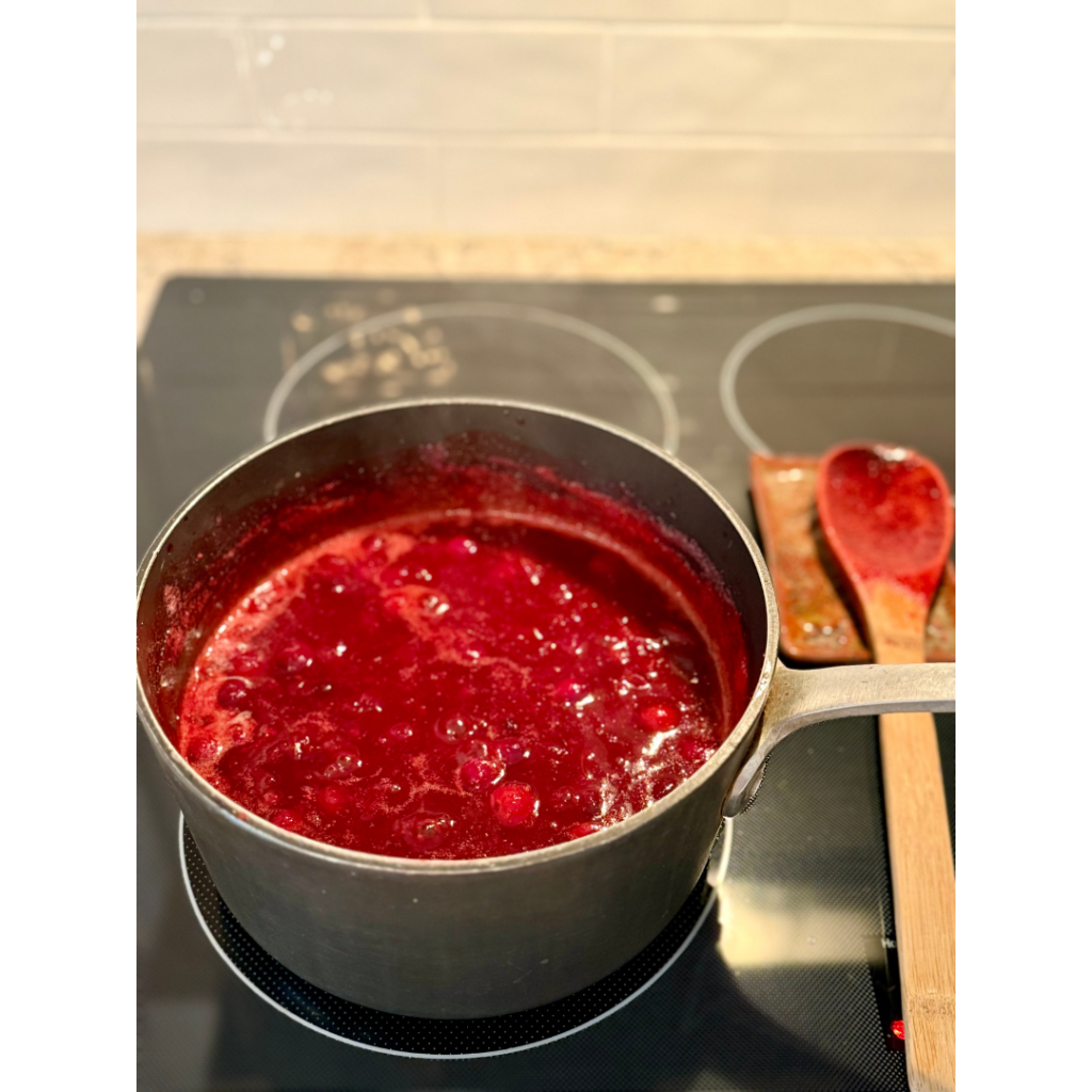 Cranberries in medium saucepan next to wooden spoon on stove top showing cranberry sauce thickening for best cranberry sauce ever recipe.