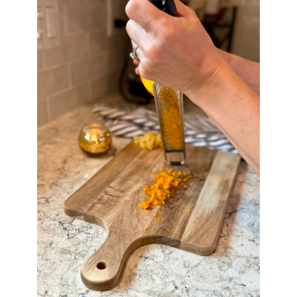 Woman using fine grater to zest orange on wooden cutting board on grey and white quartz countertop.