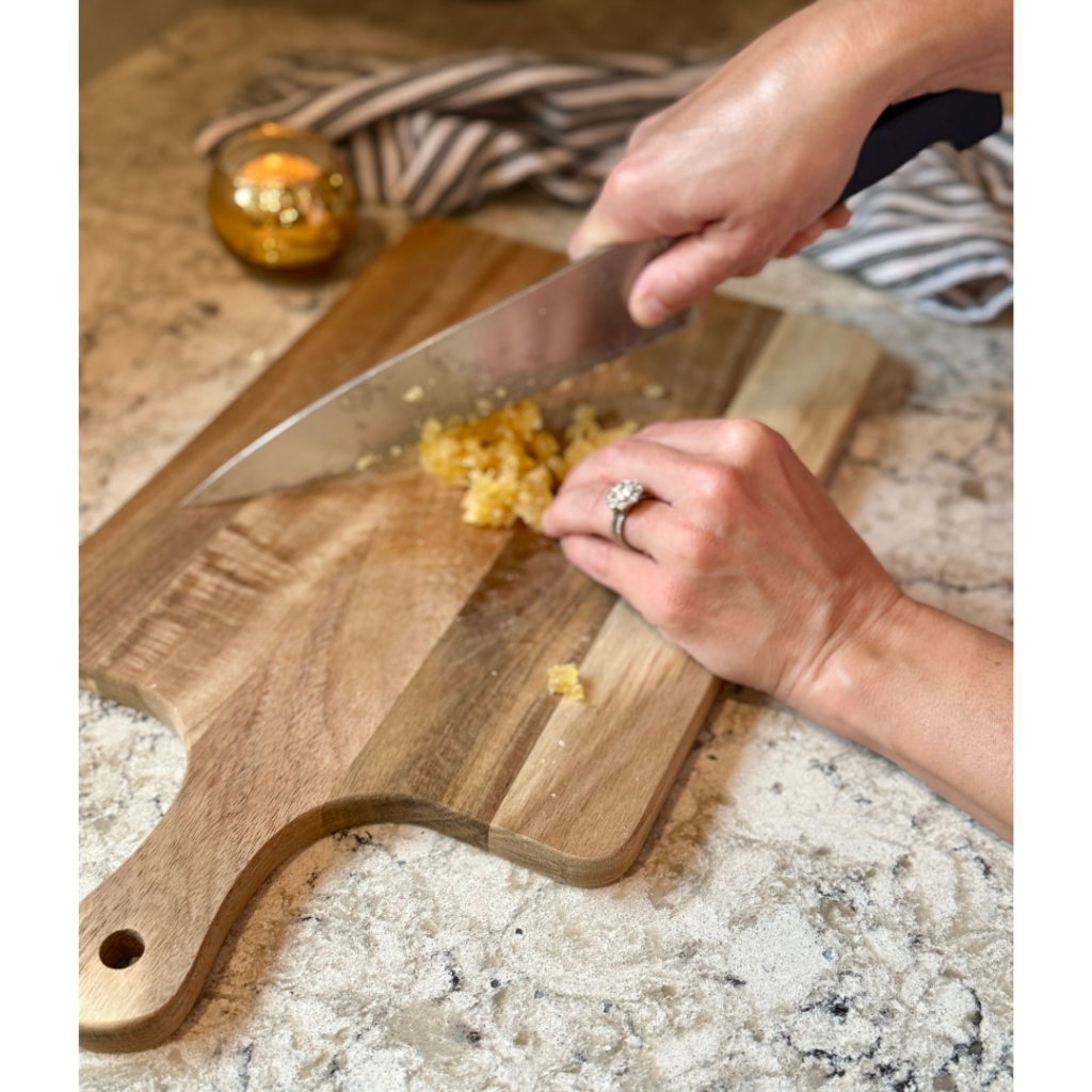 Woman cutting crystalized ginger with large knife on wooden cutting board on white and grey quartz counterop.