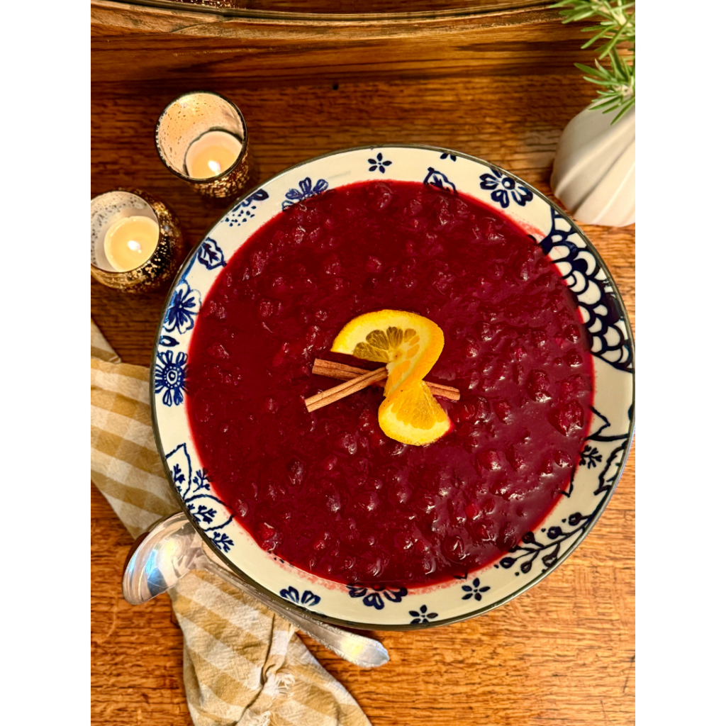 Bowl of cranberry sauce that is garnished with cinnamon sticks and orange peel, place on a wooden hutch with candles and cloth napkin.