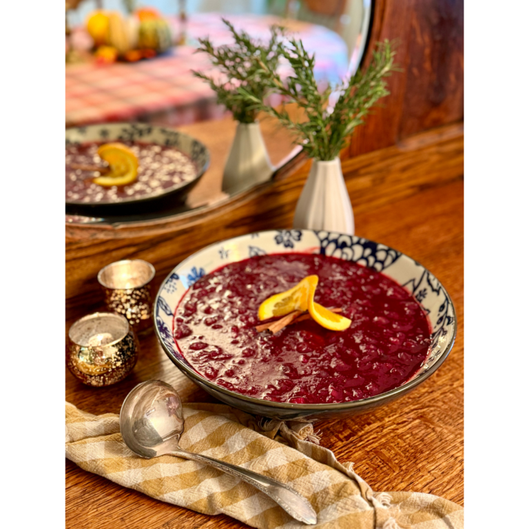 Bowl of cranberry sauce on wooden hutch with vase of rosemary and gingham cloth napkin and serving spoon.