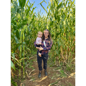 Picture of woman wearing a plaid shirt and black pants holding a baby girl in a corn maze.