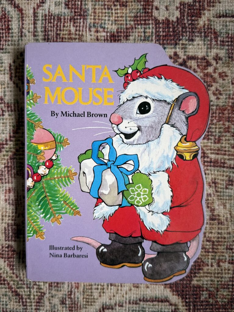 Picture of book called "Santa Mouse" and a mouse in a santa suit is on the cover. Book is placed on a multi-color red rug.