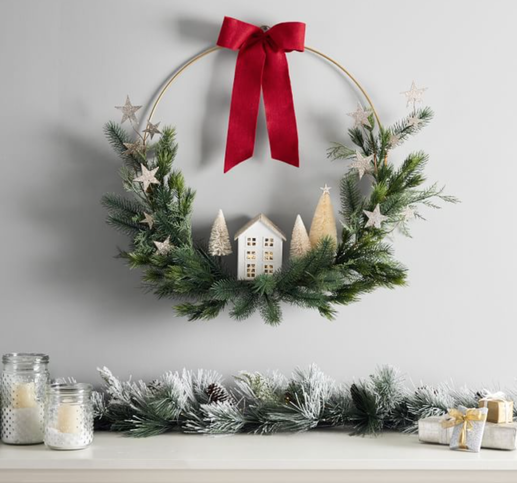 Image of Christmas Village Houses on sale in wreath that has stars and red ribbon. Wreath is hanging about a mantel with garland, white candles, and presents.