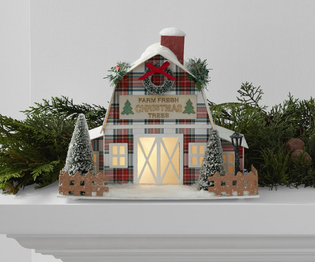 Image of Pottery Barn house that is plaid with sign that says "Farm Fresh Christmas Trees" and covered in red and green plaid. Trees and fence are in front of house with windows lit up. House is on mantel surrounded by garland. 