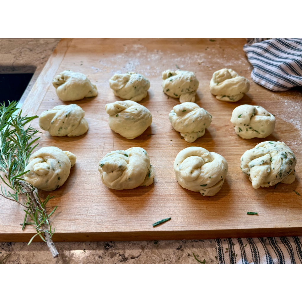 Image of 12 rolls tied in knows on lightly floured wooden cutting board, surrounded by rosemary and black and white stripped dish cloth. 