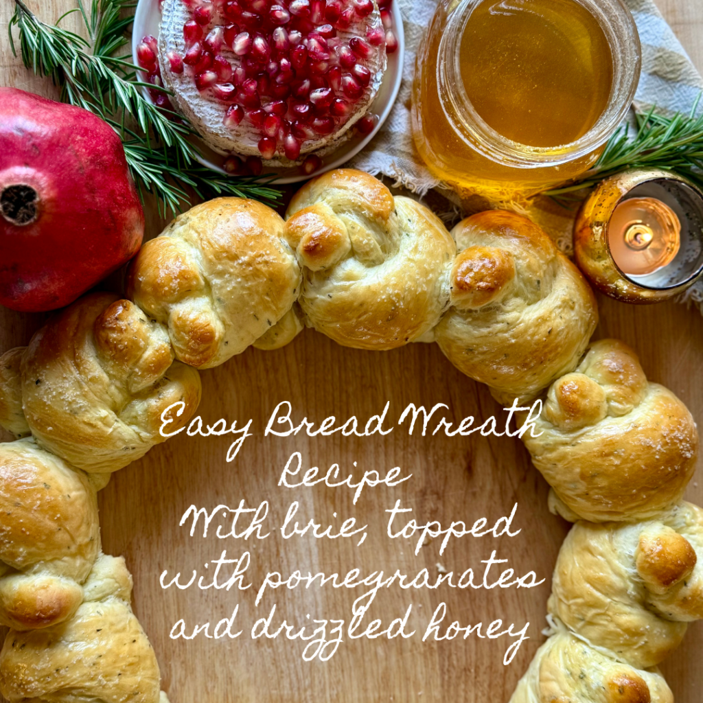 Image with cooked bread wreath and surrounded by a lit candle in a gold holder, honey in a glass jar, a small plate with pomegranates, rosemary, and a whole pomegranate. Text says "easy bread wreath recipe with brie, topped with pomegranates, and drizzled honey. 