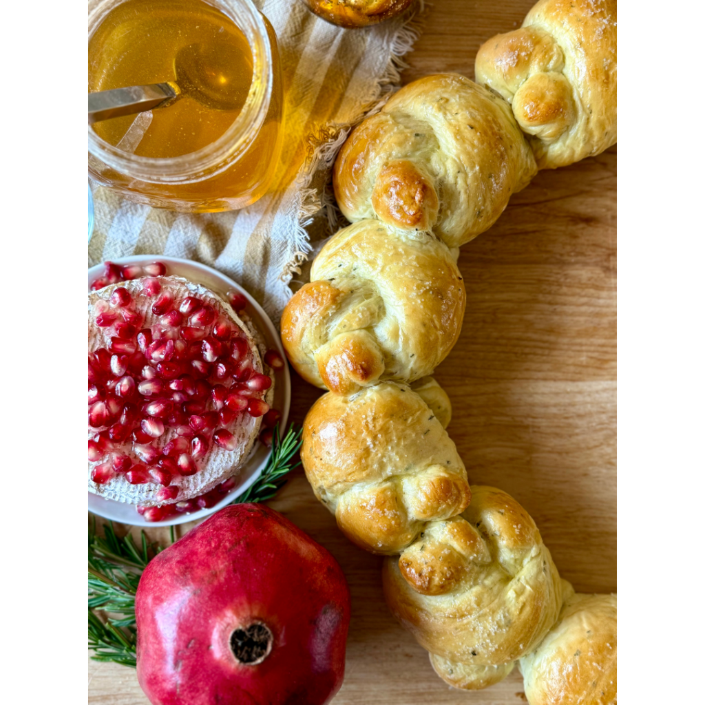 Image of bread rolls on wooden cutting board, surrounding by pomegranate, rosemary sprigs, wheel of brie with pomegranates, a jar of honey with a spoon on a gingham cloth.