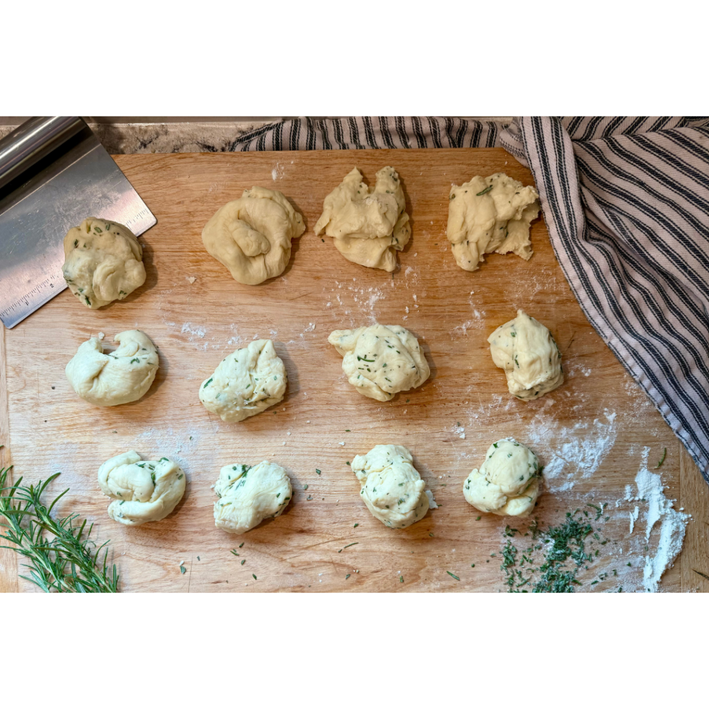 Image of dough separated into 12 pieces with rosemary sprinkled in on a wooden cutting board with a bench knife and black and white stripped dish cloth next to it. 