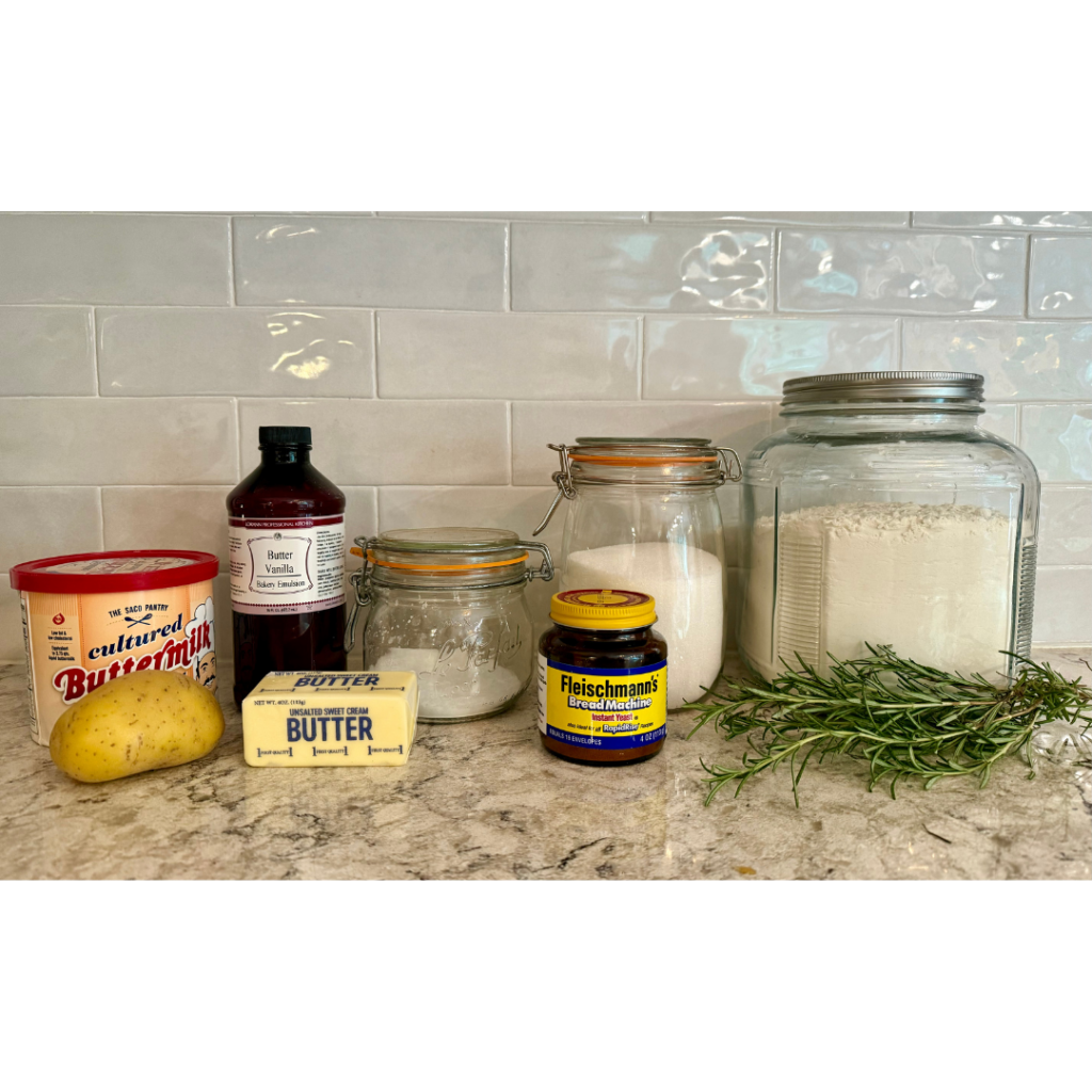 Image of ingredients (flour, rosemary, sugar, salt, yeast, butter, buttery dough enhancer, powdered buttermilk, and Yukon potato) for easy bread wreath recipe on grey, brown and white quartz counter with white subway tile background. 