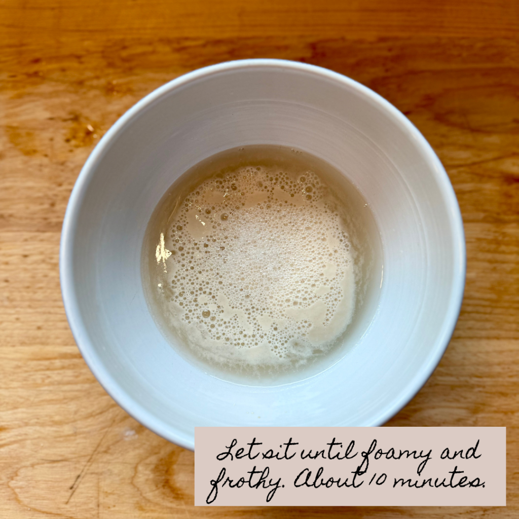 Image of foamy yeast mixture in white bowl on wooden cutting board and text that says "let sit until foamy and frothy. About 10 minutes. "