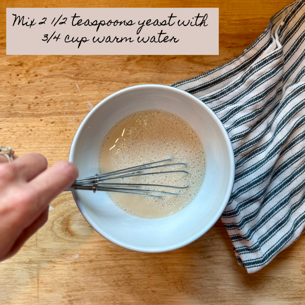 Image of woman's hand stirring yeast and water with whisk in bowl on wooden cutting board next to black and white stripped dish towel. Text says "mix 2 1/2 teaspoons yeast with 3/4 cup warm water.)