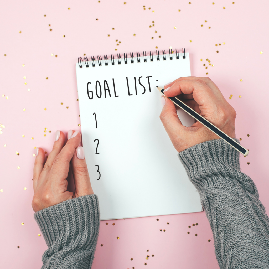 Image of woman wearing a grey sweater writing a goal list on a notebook with a pink background with gold confetti.