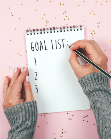 Image of woman wearing a grey sweater writing a goal list on a notebook with a pink background with gold confetti.