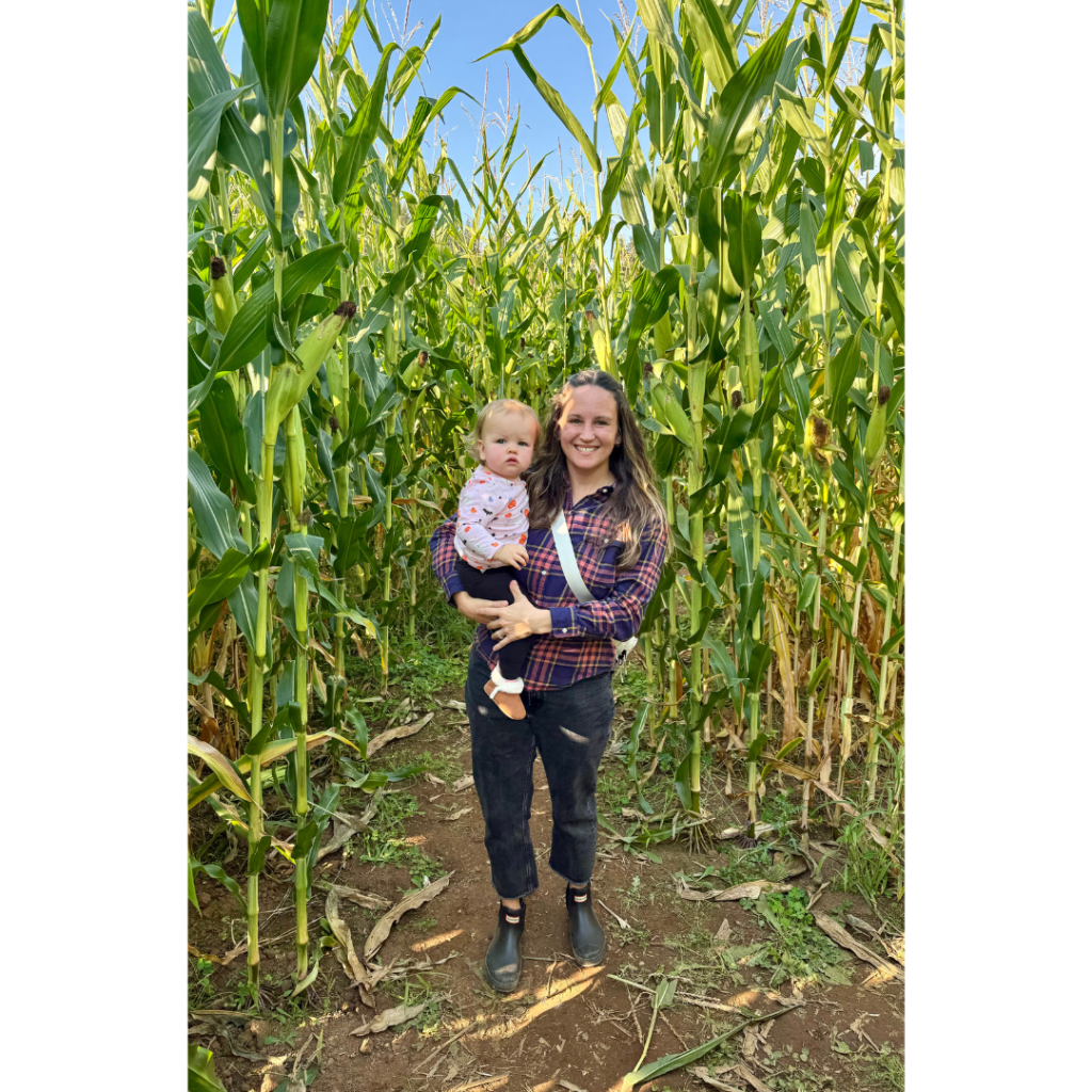 About Dana picture. Picture of woman in plaid shirt and black pants holding girl toddler in a pink shirt in corn field. 