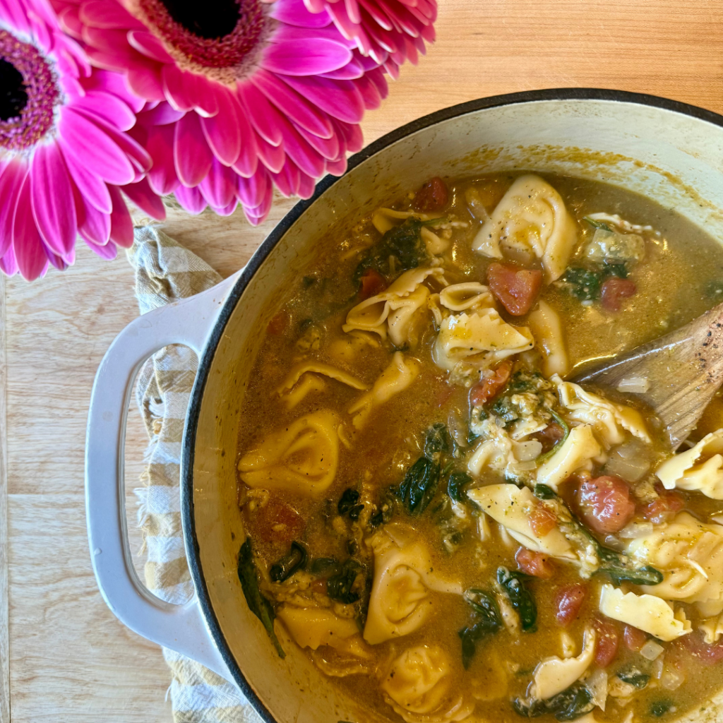 easy tortellini soup. Image of white enamel dutch oven with tortellini soup that has tomatoes and spinach. Pot sitting next to pink flower. 