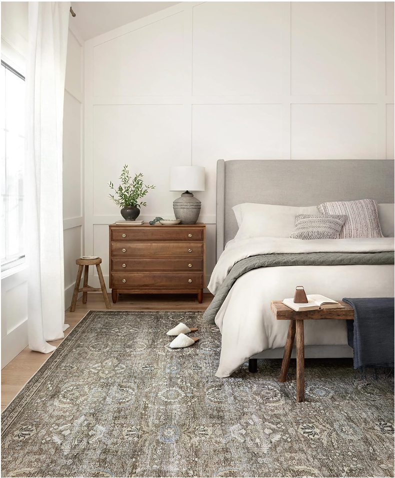 Everyday household essentials. Image of bedroom with sage green vintage rug with bed that has a grey headboard and white bedding next to a dark brown wooden night stand that has a grey ceramic lamp and plant. 