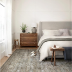 Everyday household essentials. Image of bedroom with sage green vintage rug with bed that has a grey headboard and white bedding next to a dark brown wooden night stand that has a grey ceramic lamp and plant.