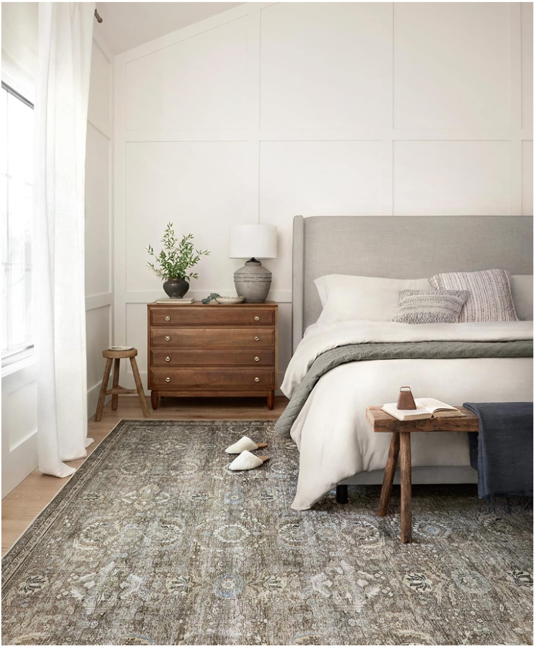 Everyday household essentials. Image of bedroom with sage green vintage rug with bed that has a grey headboard and white bedding next to a dark brown wooden night stand that has a grey ceramic lamp and plant.