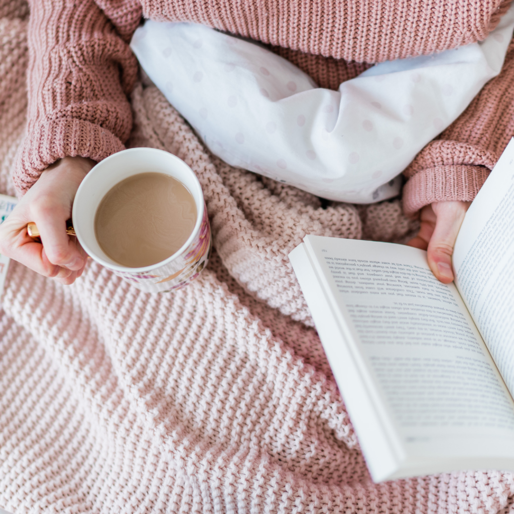 Image of woman reading a book in bed with a cup of tea. Woman is wearing a pink knit sweater with a pink knit blanket.