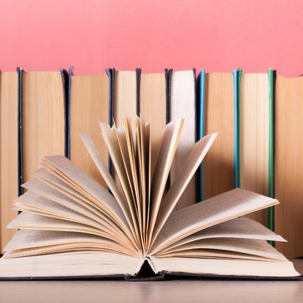 Image of books lined up, pages facing out and one book open with pages fanned open. Background above the books is pink. 