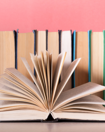 Image of books lined up, pages facing out and one book open with pages fanned open. Background above the books is pink.