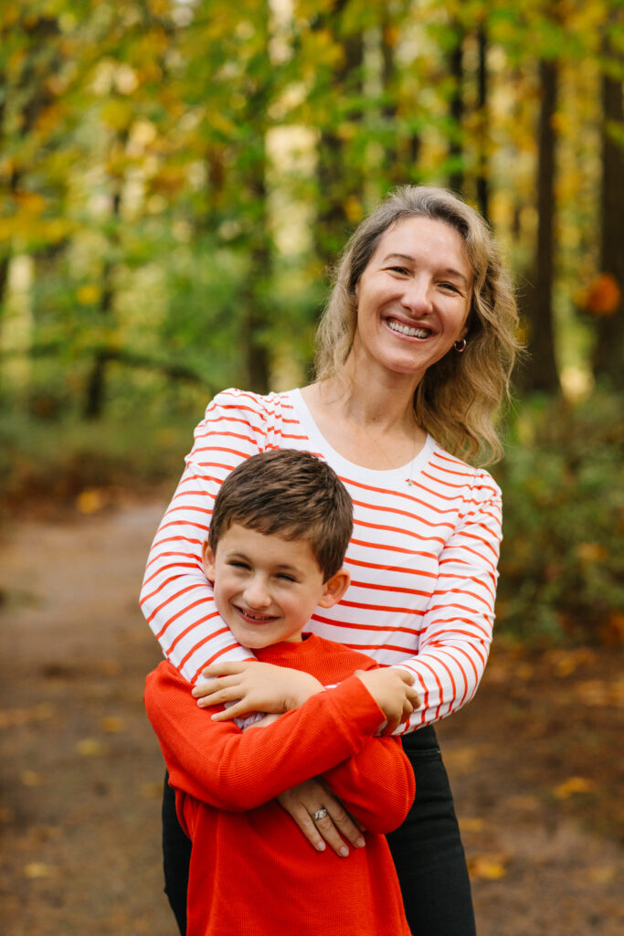 Mothering. Image of mother and son in forest smiling. Mother has blond hair and white and red striped long sleeve shirt and son is wearing a red shirt.