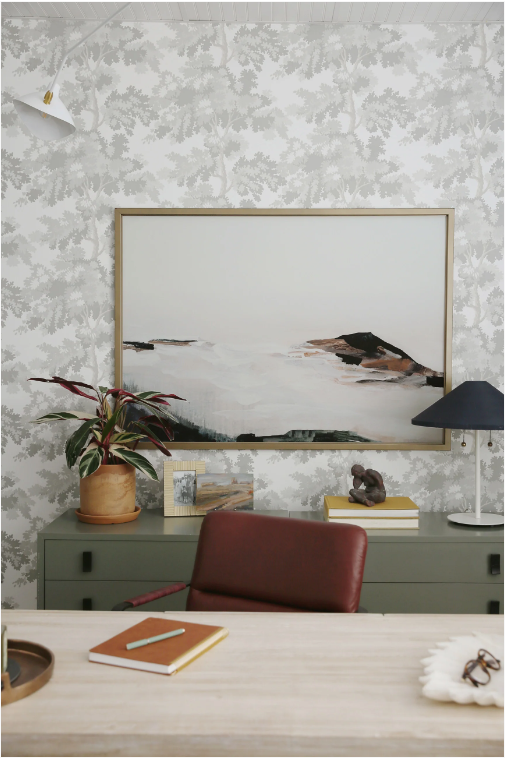 Image of art print of abstract mountain top with clouds in front of it and sky. Painting is hanging on wallpaper wall with green dresser below with plant, books, and lamp on it. In the front of the image is a desk with a red chair.