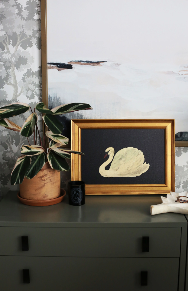 Affordable art prints. Image of art of swan in gold frame leaning against abstract art that is hanging on a wallpaper wall. Swan painting is sitting on a green dresser next to a plant and black candle.