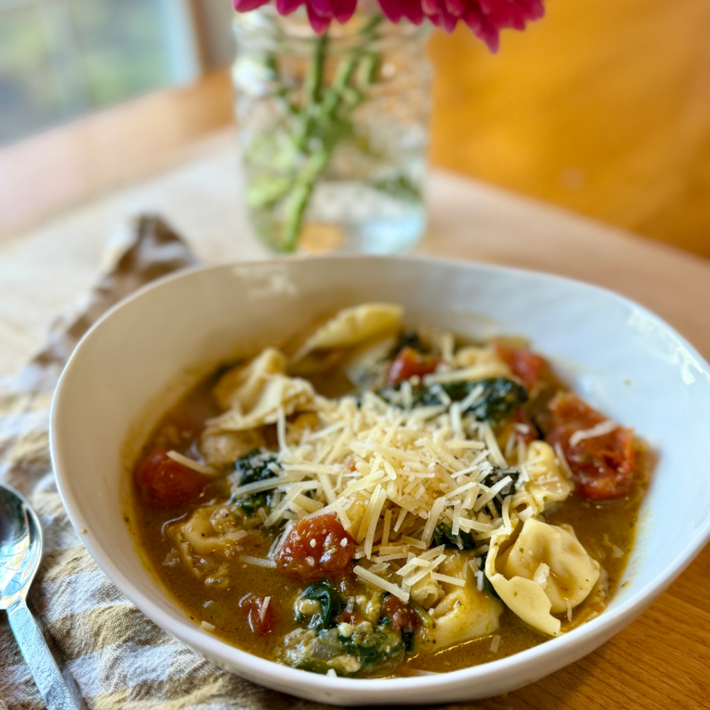 easy tortellini soup with spinach. Image of bowl of tortellini soup with spinach, tomato, and topped with parmesan. Tan and white gingham cloth napkin next to bowl of soup. 