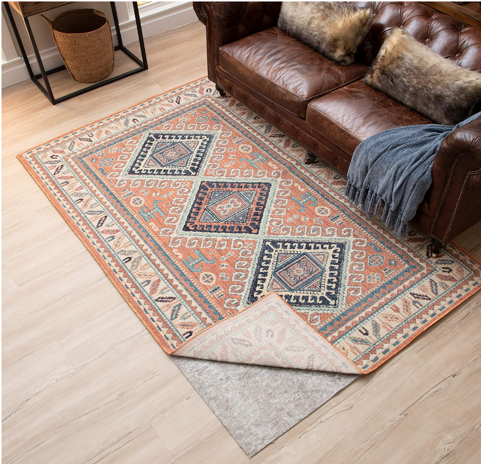 household essentials list items. Image of area rug with oranges, blues, and greens in an aztec pattern that is folded back to show rug pad. There is a dark brown leather couch with faux fur pillows and blur throw blanket and a wicker basket in the corner next to a side table. 