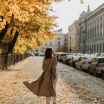 Inspiring podcasts. Image of woman in tan trench coat walking down car lined street in the city with yellow leaves on trees.