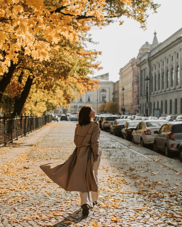 Inspiring podcasts. Image of woman in tan trench coat walking down car lined street in the city with yellow leaves on trees.