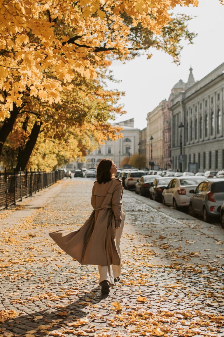 Inspiring podcasts. Image of woman in tan trench coat walking down car lined street in the city with yellow leaves on trees.