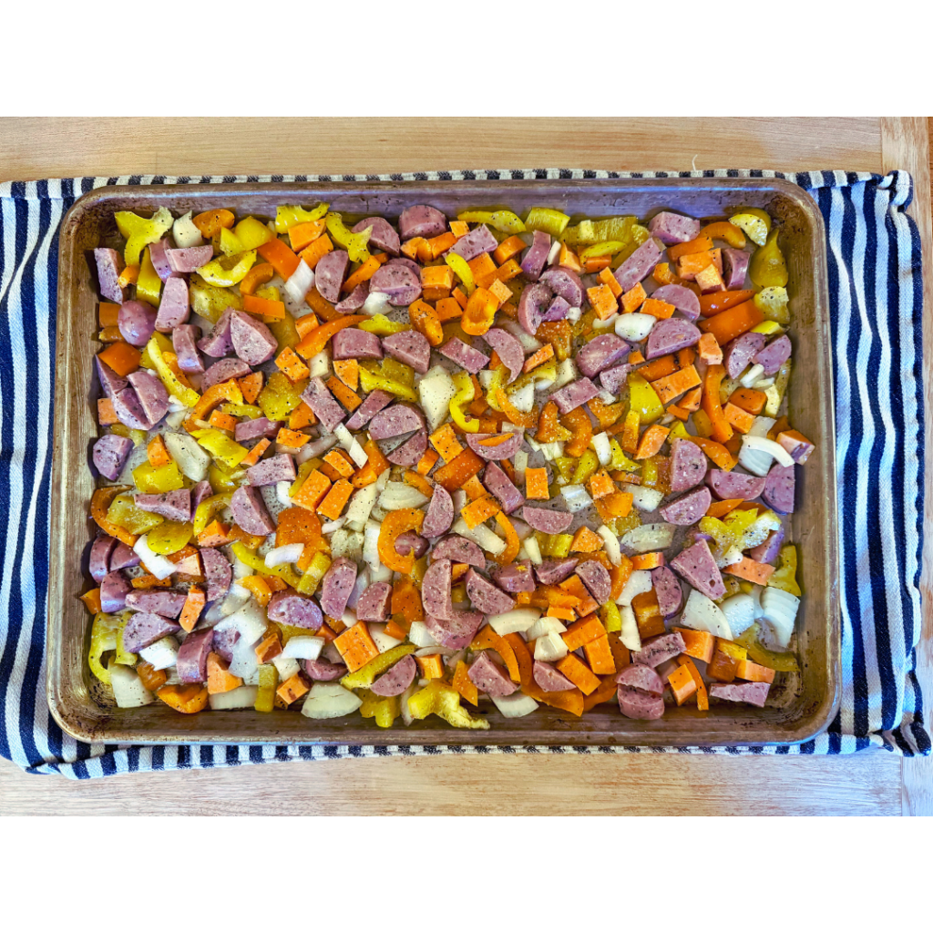 easy sheet pan chicken and veggies. Image of chopped up sweet potato, yellow bell pepper, chicken sausage, and white onion on a sheet pan that's set on a black and white striped dish towel on a wood counter top. 
