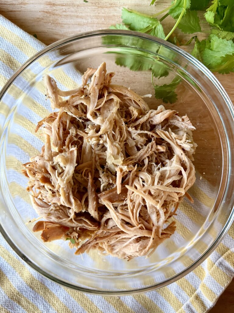 crockpot shredded chicken. Image of shredded chicken in clear glass bowl on wooden cutting board with yellow and white stripped cloth napkin, next to cilantro. 