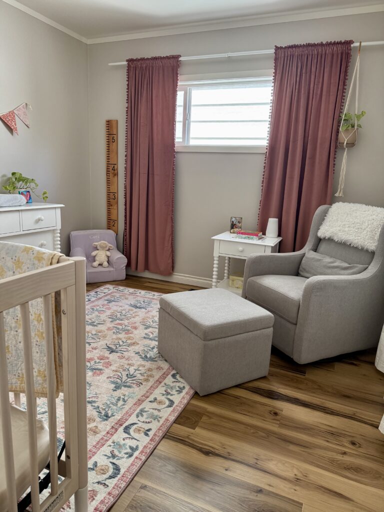 baby girl nursery tour. Image of girl nursery with crib, floral rug, grey glider, pink curtains, growth chart ruler, and white dresser.
