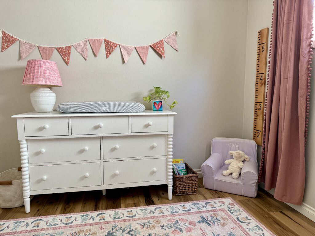 baby girl nursery design. Image of white dresser with lamp, changing pad, plant, and art on top and fabric bunting hanging on top. Growth chart ruler hanging in the corner of the room with a lavender chair and pink curtains.