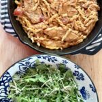 easy chicken recipe. Image of creamy bruschetta chicken in cast iron skillet on top of black and white cloth napkin on wood countertop next to arugula salad in blue and white patterned bowl.