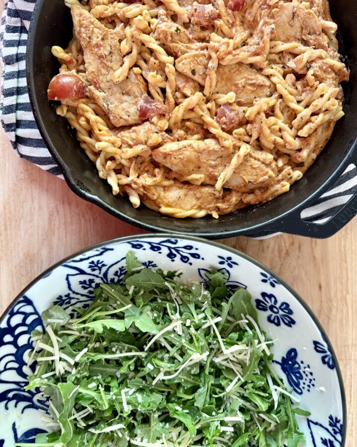 easy chicken recipe. Image of creamy bruschetta chicken in cast iron skillet on top of black and white cloth napkin on wood countertop next to arugula salad in blue and white patterned bowl.