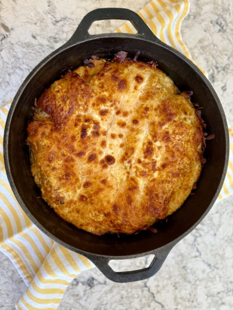 Image of cooked dutch oven lasagna in cast iron dutch oven on yellow and white striped towel on a grey and white quartz countertop. 