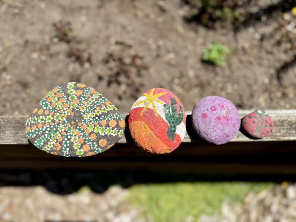 fun art projects for kids. Picture of 4 rocks lined up on the end of a garden bed. Rocks are painted in a variety of colors and patterns.