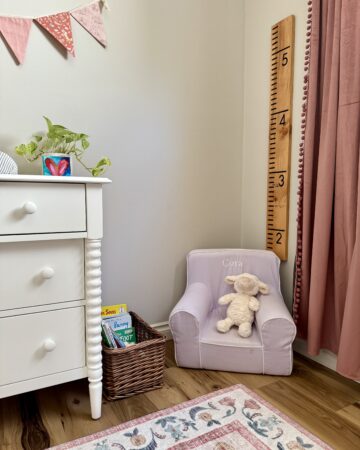 how to make a growth chart ruler. image of girl's nursery with pink velvet curtains, lilac chair that says "Cora" with books in wicker basket next to it white dresser with plant and heart painting on top with fabric bunting hanging above and in front of flower print rug.