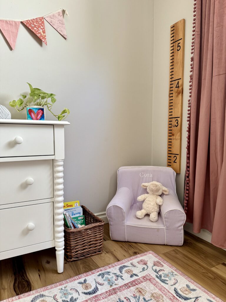 baby girl nursery room tour. Image of white dresser with plant and heart painting on canvas next to wicker basket with books, lilac purple chair, wooden growth chart ruler, and pink velvet curtains in front of floral pattern rug.