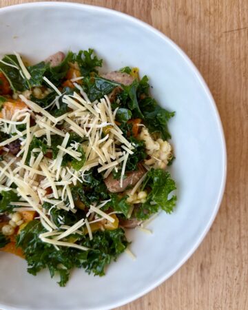 easy sheet pan sausage and veggies. Image of veggies, kale, grains, and parmesan in white ceramic bowl on wood counter.