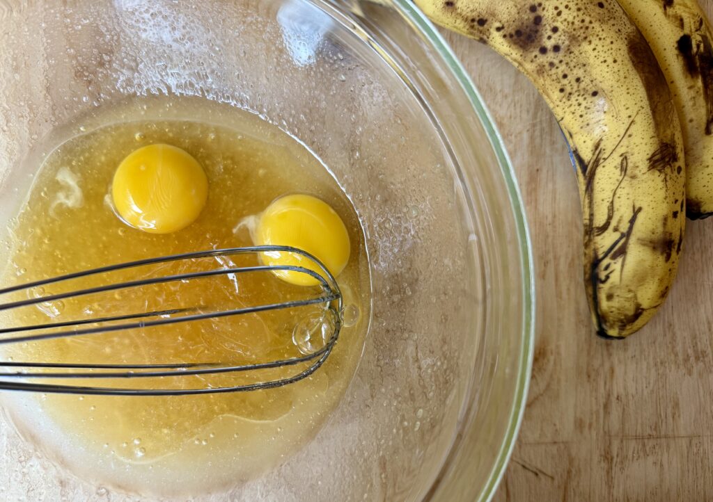 easy sourdough banana bread. image of coconut oil, honey, and eggs in glass bowl with whisk next to bananas on wooden counter.