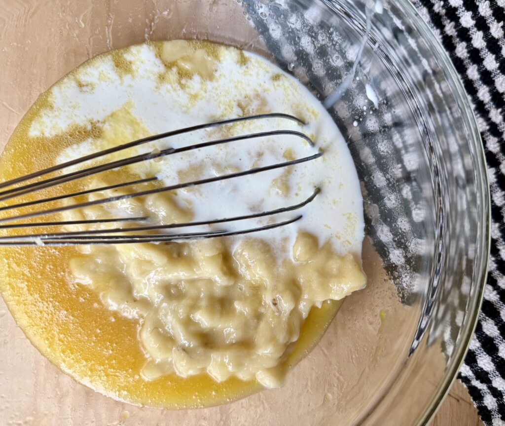 best banana bread recipe ingredients. Image of coconut oil, honey, milk, and mashed bananas in glass bowl next to black and white dish cloth on wood counter top.